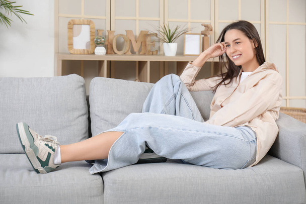 Mujer joven y bonita acostada en un sofá gris en la sala de estar - Foto, Imagen