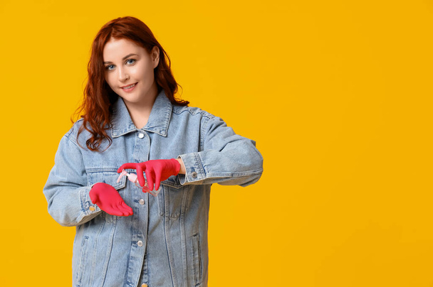 Beautiful young happy woman in rubber gloves applying sanitizer on yellow background - Photo, Image