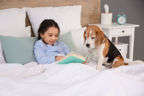Linda niña asiática con Beagle perro libro de lectura en el dormitorio - Foto, imagen