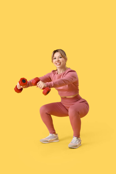 Jeune entraîneur de fitness féminin faisant de l'exercice avec des haltères sur fond jaune - Photo, image