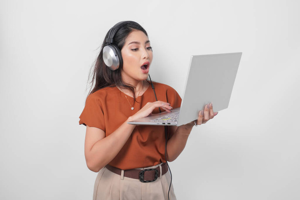 Shocked young woman wearing brown shirt and headphone while holding laptop to listen music isolated over white background. - 写真・画像