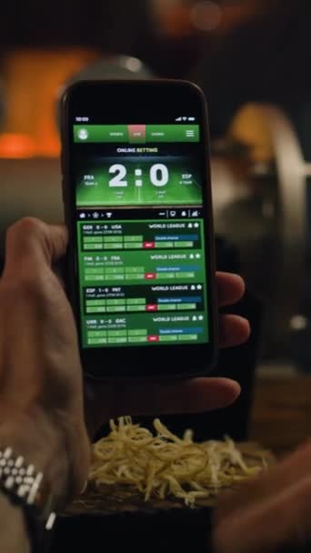 Vertical shot of sports fan watching game score and bookmaker ratings using mobile phone. Caucasian man bets on football world league match online sitting at bar counter in pub at night. Gambling - Footage, Video