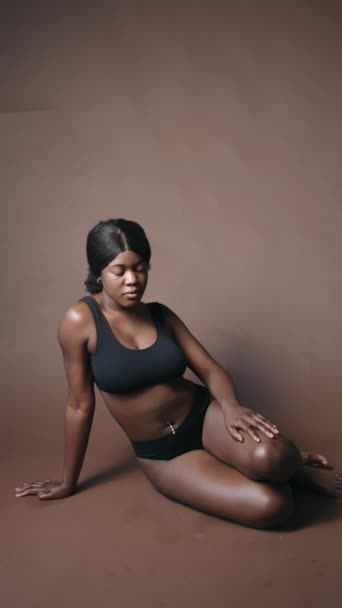 Vertical shot de sensual chica negra en sujetador y bragas sentado en el suelo en el estudio, acariciando su cuerpo y mirando a la cámara - Metraje, vídeo