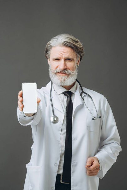 Седовласый врач в белом медицинском халате показывает экран телефона на камеру. Медицинский макет. - Фото, изображение