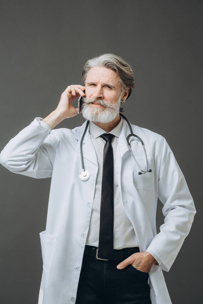 A middle-aged man with a beard and gray hair, dressed in a medical gown, is talking on the phone. Isolated on gray background. - Photo, Image