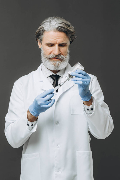 Γκρι μαλλιά ανώτερος γιατρός σε λευκό παλτό κρατώντας σύριγγα με ένεση, απομονώνονται σε γκρι φόντο. - Φωτογραφία, εικόνα