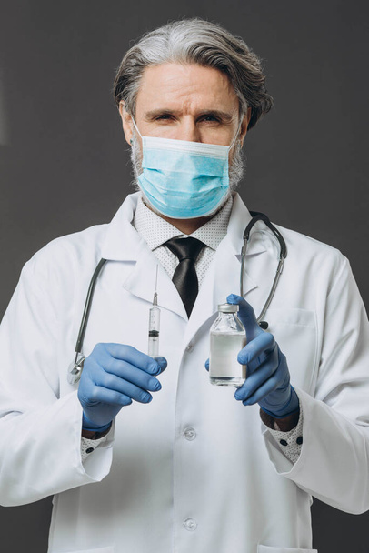 Starszy lekarz w masce ochronnej i rękawiczkach przygotowuje się do wstrzyknięcia, trzymając szczepionkę i strzykawkę.. - Zdjęcie, obraz
