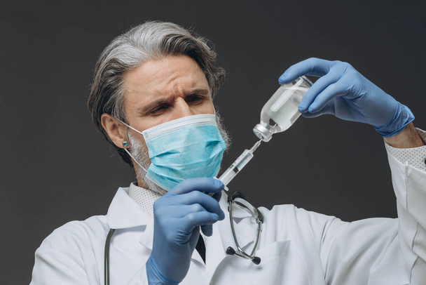 Un médico de cabecera con una máscara protectora y guantes se está preparando para una inyección, inyectando la vacuna en una jeringa. - Foto, Imagen