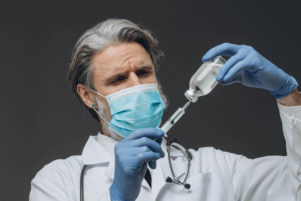 Starszy lekarz w masce ochronnej i rękawiczkach przygotowuje się do wstrzyknięcia, pobierając szczepionkę do strzykawki.. - Zdjęcie, obraz