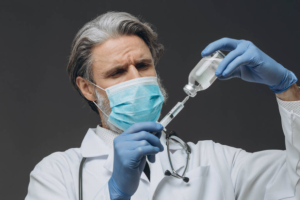 Un médecin chevronné portant un masque protecteur et des gants se prépare pour une injection, en tirant le vaccin dans une seringue. - Photo, image
