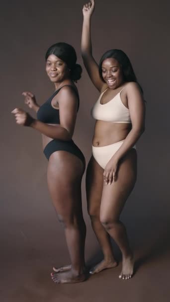 Tiro vertical de dos mujeres negras felices con hermosas curvas con ropa interior minimalista bailando y divirtiéndose en el estudio - Metraje, vídeo