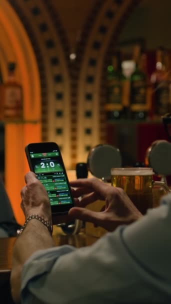 Vertical shot of fan checks game score and bookmaker ratings in app using mobile phone, talks with bartender. Caucasian man bets on football match online sitting at bar counter in pub. Gambling - Footage, Video