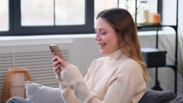 Vriendelijke blanke vrouw chatten online en lachen met behulp van de telefoon thuis op de bank. Genieten van vrije tijd en rusten online met social media. - Video