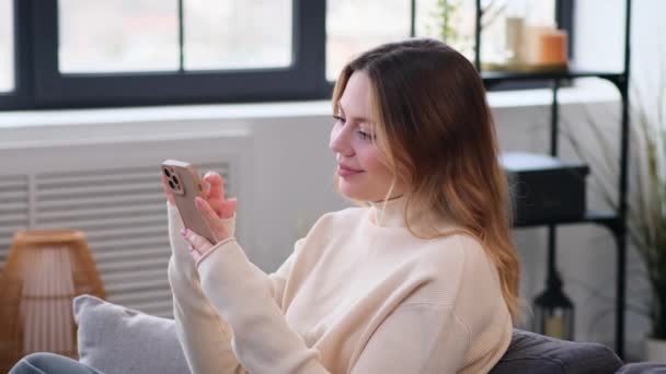 Iloinen ja onnellinen valkoihoinen nainen selaamassa internetiä älypuhelimella, rentouttava verkossa sohvalla kotona olohuoneessa. Kotimaan vapaa-aika ja eläke. - Materiaali, video