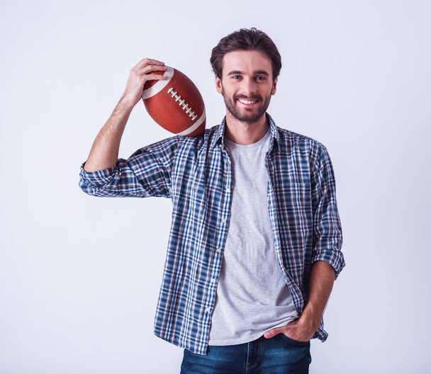 Hombre barbudo guapo en ropa casual está sosteniendo una pelota, mirando a la cámara y sonriendo, sobre un fondo claro
 - Foto, Imagen