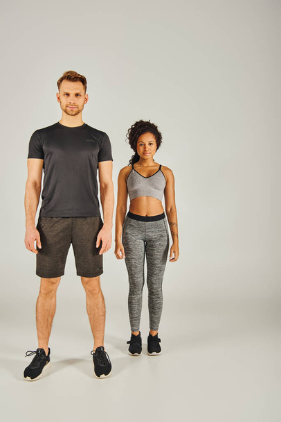 Ein junges interrassisches Sportpaar in aktiver Kleidung steht nebeneinander auf grauem Studiohintergrund. - Foto, Bild