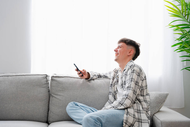 Šťastný mladý muž se směje při používání telefonu sedí na pohovce - Fotografie, Obrázek