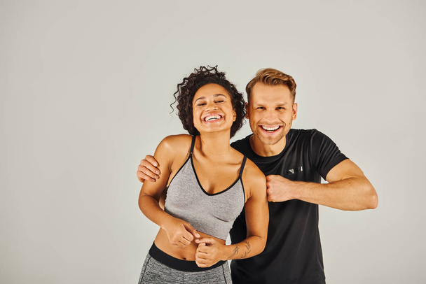 Un jeune couple sportif interracial en tenue active posant en toute confiance pour la caméra dans un studio sur fond gris. - Photo, image