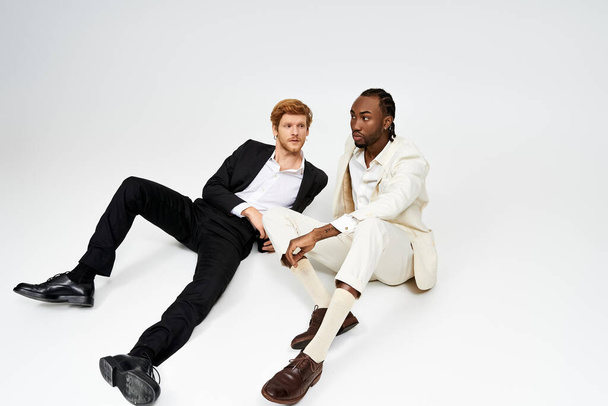 Deux beaux hommes multiculturels en tenue élégante assis les uns à côté des autres sur un fond blanc. - Photo, image