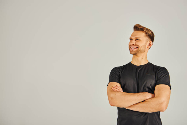 Un joven atlético con una camiseta negra posa confiadamente con los brazos cruzados en un estudio sobre un fondo gris. - Foto, imagen