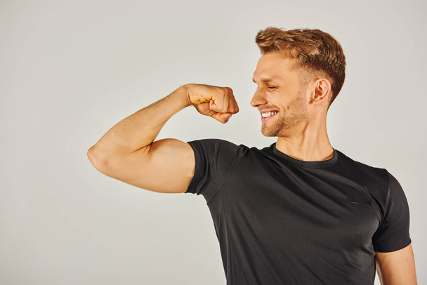 Um jovem atlético em desgaste ativo flexiona seu bíceps com confiança contra um fundo cinza neutro. - Foto, Imagem