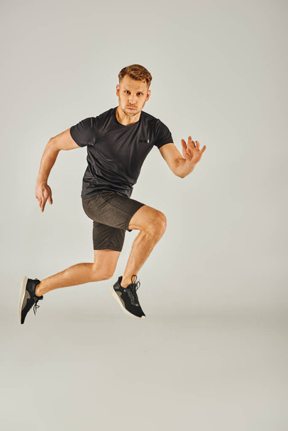 Młody sportowiec w czarnej koszulce energicznie skacze w studiu z szarym tłem. - Zdjęcie, obraz