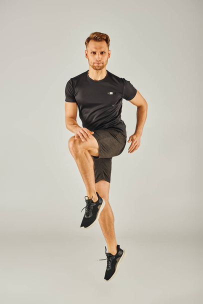 Nuori urheilullinen mies mustassa t-paidassa ja shortseissa hyppää energisesti harmaalla taustalla studioympäristössä. - Valokuva, kuva