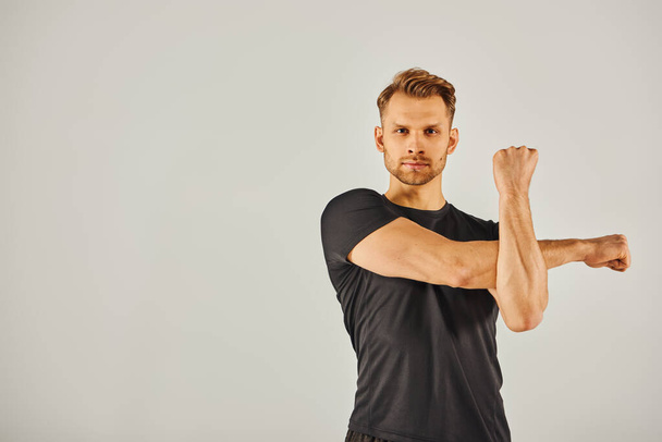 A young athletic man in active wear flexes his arm against a gray background in a dynamic display of strength and fitness. - Photo, Image