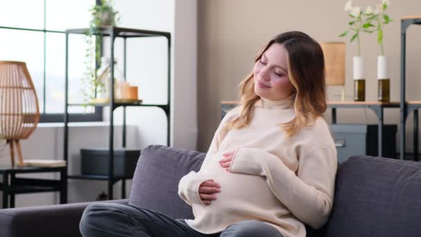 Gelukkig blanke jonge zwangere vrouw raakt buik op de bank thuis woonkamer. Thuis rusten, bevalling en moederschap verwachten. - Video