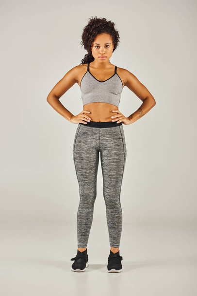 Ajuste mujer afroamericana en leggings grises con estilo posa con confianza contra un fondo gris en un entorno de estudio. - Foto, imagen