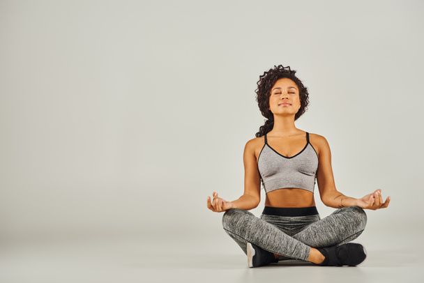 A young African American woman in activewear meditates peacefully on a gray background in a studio setting. - Photo, Image