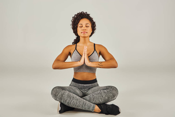 Nuori afroamerikkalainen nainen aktiivisessa käytössä meditoi rauhallisesti harmaalla pohjalla studioympäristössä. - Valokuva, kuva