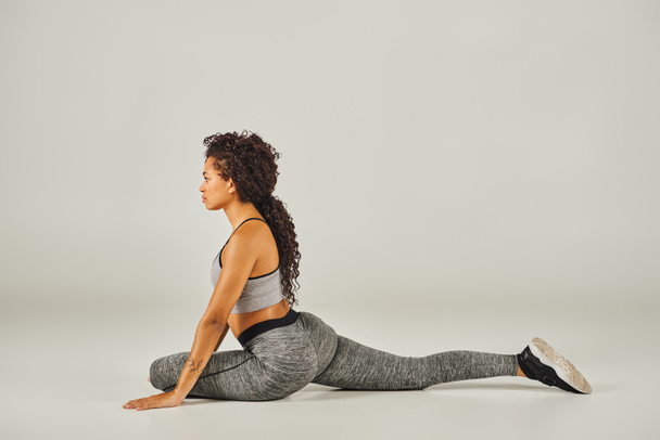 A young African American woman in active wear gracefully stretches in a squat position on a grey background. - Photo, Image
