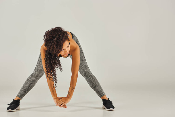Eine junge Afroamerikanerin in aktiver Kleidung streckt anmutig ihre Beine vor grauem Hintergrund in einem Atelier. - Foto, Bild