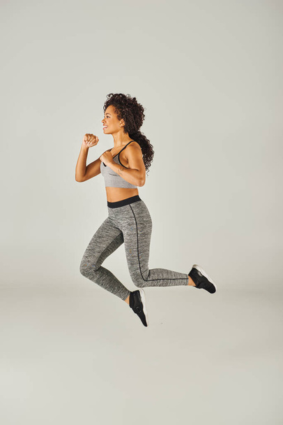 Eine junge Afroamerikanerin macht einen dynamischen Sprung in einem grauen Sport-BH und Leggings in einem Studio-Setting. - Foto, Bild
