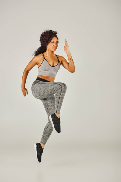 Uma jovem mulher afro-americana em um sutiã esportivo e leggings salta poderosamente em um fundo de estúdio cinza. - Foto, Imagem