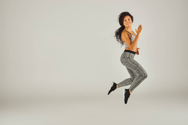 アスレチックな服装の若いアフリカ系アメリカ人女性は,スタジオの設定で明白な灰色の背景に喜んでジャンプします. - 写真・画像