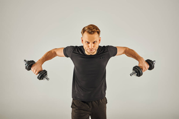 Un joven deportista en ropa activa sostiene dos pesas en un estudio con un fondo gris. - Foto, imagen