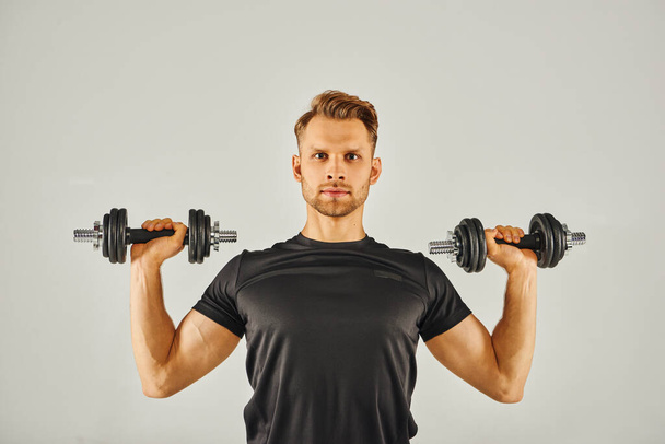 Молодий спортсмен у активному вбранні тримає двох гантелів у студії на сірому фоні, зосереджуючись на фізичній підготовці та силі. - Фото, зображення