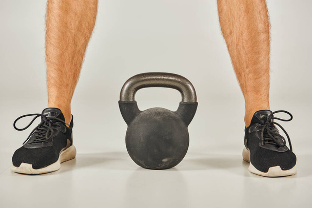 Young sportsman in active wear standing next to a kettlebell in a studio with a grey background. - Photo, Image