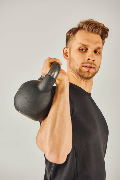 Un jeune sportif en tenue active montrant sa force en tenant une kettlebell sur son bras sur un fond de studio gris. - Photo, image