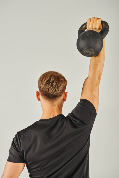 Un jeune sportif en tenue active soulève intensément une kettlebell dans un studio au fond gris. - Photo, image
