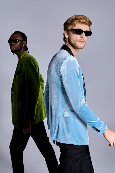 Due uomini diversi in abiti eleganti camminano con fiducia in una strada urbana indossando occhiali da sole. - Foto, immagini
