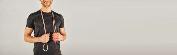 Молодий спортсмен в активному одязі посміхається, тримаючи мотузку в студії з сірим фоном. - Фото, зображення