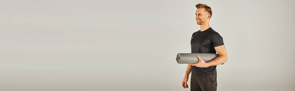Young sportsman in active wear holding a yoga mat in front of a grey background. - Photo, Image