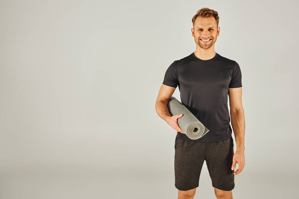 Mladý muž v aktivním oblečení drží podložku na jógu před šedým pozadím. - Fotografie, Obrázek