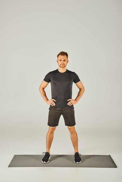 Ένας νεαρός αθλητής σε ενεργό φθορά στέκεται σε ένα χαλί μπροστά από ένα λευκό φόντο, επιδεικνύοντας ισορροπία και εστίαση σε ένα στούντιο. - Φωτογραφία, εικόνα