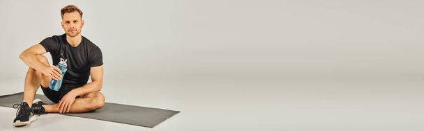 Nuori aktiiviseen kulumiseen pukeutunut urheilija keskittyy hiomaan kyykky-asentoaan istuessaan matolla studiossa, jolla on harmaa tausta. - Valokuva, kuva