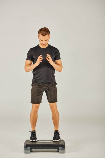 Ein junger Sportler in aktiver Kleidung fordert sein Gleichgewicht auf einem Stepper in einem Studio mit grauem Hintergrund heraus. - Foto, Bild