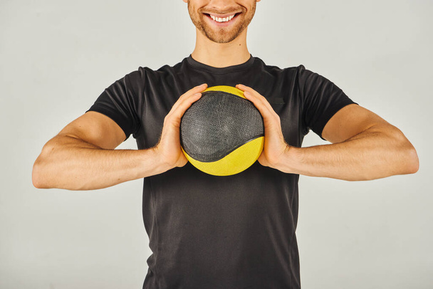 Молодой спортсмен в активной одежде энергично держит яркий желтый и черный шар в студии на сером фоне. - Фото, изображение